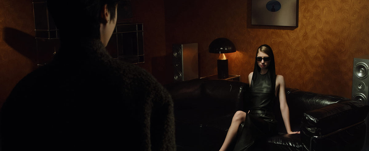 Woman seated on black leather sofa, warm amber lighting creates a cinematic and mysterious mood.